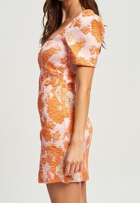 Femme portant une robe ajustée, texturée, avec de courtes manches bouffantes, dans un motif floral orange, blanc et beige, se tenant en profil latéral.
