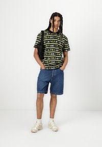 Grafisk svart t-skjorte med fargerike mønstre, sammen med lyseblå denimshorts og beige sneakers. Tilbehør: svart crossbody-veske.