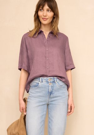 Kvinde iført en løs lilla skjorte med knapper og lyseblå højtaljede jeans, der holder en beige jakke i den ene hånd mod en beige baggrund.