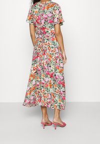 Femme portant une robe midi florale avec des motifs roses, orange et verts, associée à des sandales à talons roses, se tenant devant un fond blanc.
