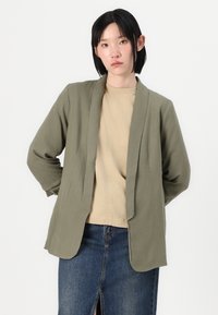 Oliven grøn blazer i blødt stof med een afslappet pasform, der har lange ærmer og åben front, parret med en beige top og et denim nederdel.