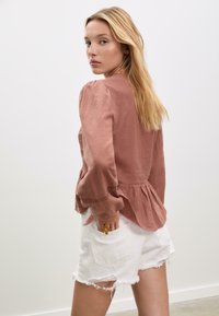 Brun texturerad blus med puffärmar och peplumslut, kombinerad med vita slitna denimshorts. Modellen är vänd lite åt sidan, med handen i fickan.