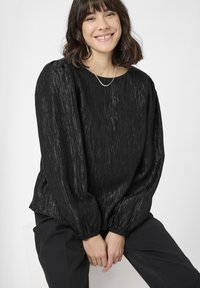 Bluză neagră texturată cu o croială lejeră, mâneci balon și un decolteu larg. Pur și simplu cu pantaloni negri, care au un luciu subtil.