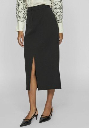 Falda de lápiz negra con una abertura frontal, diseño ajustado y textura suave, combinada con tacones negros de punta afilada.