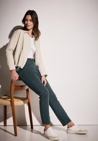 Chaqueta cremosa con cremallera, camiseta blanca, pantalones verde oscuro y zapatillas blancas. La silla tiene un asiento trenzado y un marco de madera.