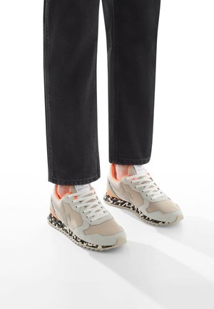 Sneaker beige e grigie con tomaia in mesh, lacci bianchi e suola a motivo bianco e nero. Accompagnati da pantaloni neri a gamba dritta.