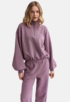 Kobieta w mauve dresie z półzamkiem i spodniach z elastyczną talią na jednolitym jasnym tle.