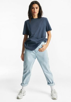 Kobieta stojąca na białym tle, ubrana w ciemnoniebieską koszulkę, jasno niebieskie podwinięte dżinsy, białe skarpetki i białe trampki.