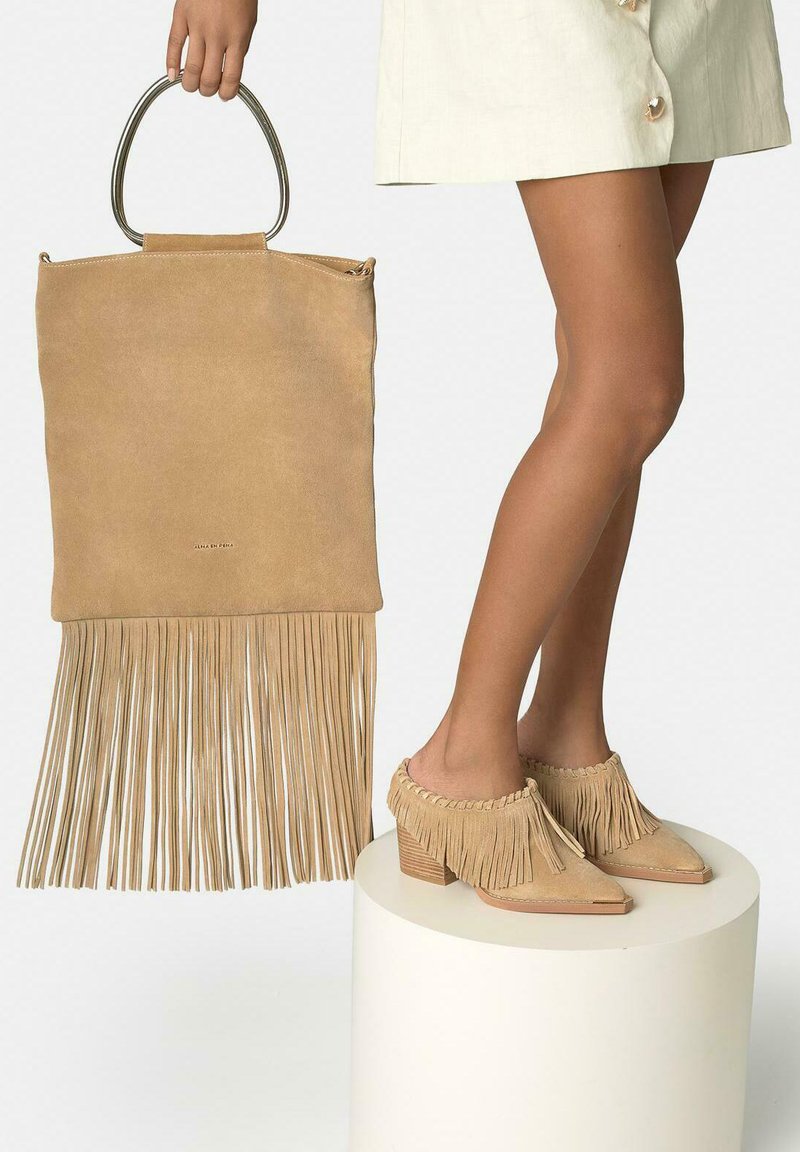 Beżowa zamszowa torebka z frędzlami i metalowymi rączkami trzymana jedną ręką, zestawiona z beżowymi zamszowymi butami na słupku z frędzlami, noszonymi na nogach ubranych w kremową spódnicę.