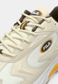 Zapatillas de cuero beige con acentos crema, con cordones marrones, un logo estilizado y paneles laterales texturizados. Suela de goma con detalle amarillo.