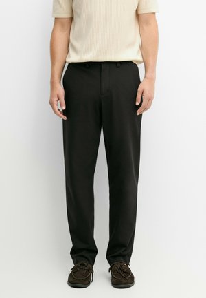 Uomo che indossa pantaloni neri a gamba dritta, maglietta beige a maniche corte a coste e scarpe stringate marrone scuro, in piedi davanti a uno sfondo semplice.