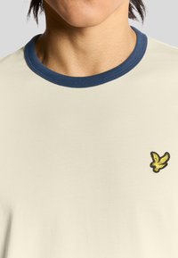Creme t-shirt med en marineblå ribberet rund hals. Har et lille gult og sort broderet fuglelogo på venstre bryst. Blød tekstur.