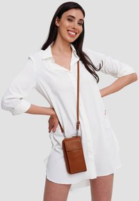 Braune Leder-Handy-Crossbody-Tasche mit strukturierter Oberfläche, ausgestattet mit einem abnehmbaren Tragegurt und einer Fronttasche. Über einem weißen Hemd getragen.