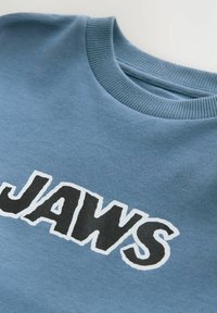 Sudadera azul con cuello redondo y la palabra "JAWS" en grandes letras negras en negrita contorneadas en blanco. Hecha de material suave.