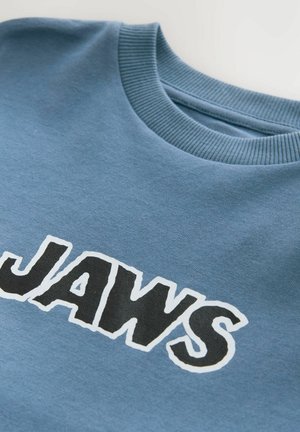 Sininen huppari, jossa on pyöreä kaula-aukko ja sana "JAWS" suurilla, lihavilla mustilla kirjaimilla, jotka on ääriviivattu valkoisella. Valmistettu pehmeästä materiaalista.