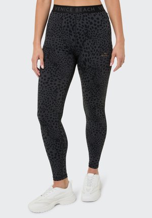 Woman wearing black leggings with dark spotted pattern, Venice Beach logo on waistband and thigh, paired with white sneakers.