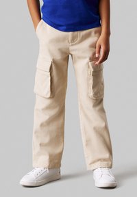 Pantalons cargo beige en tissu durable, dotés de grandes poches latérales, d'une coupe droite, et associés à des baskets blanches.