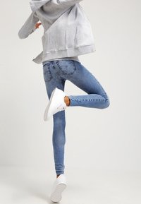 Vaqueros skinny de mezclilla en azul desgastado con una textura suave, que presentan bolsillos traseros y detalles de costura sutil, combinados con zapatillas blancas.