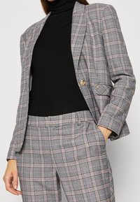 Szara i różowa kraciasta marynarka z dopasowanym blezerem i pasującymi spodniami, wykonane z teksturowanej tkaniny z pojedynczym złotym guzikiem jako akcentem. Pod spodem czarny golf.