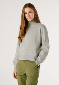 Sudadera gris con un cuello alto y puños acanalados, con un diseño corto. Combinada con pantalones verde oliva, que muestran bolsillos.