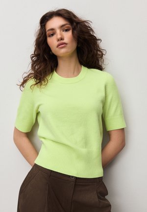 Fiatal nő göndör hajjal, világoszöld rövid ujjú pulóvert és sötétbarna nadrágot viselve, egy sík fal előtt állva.