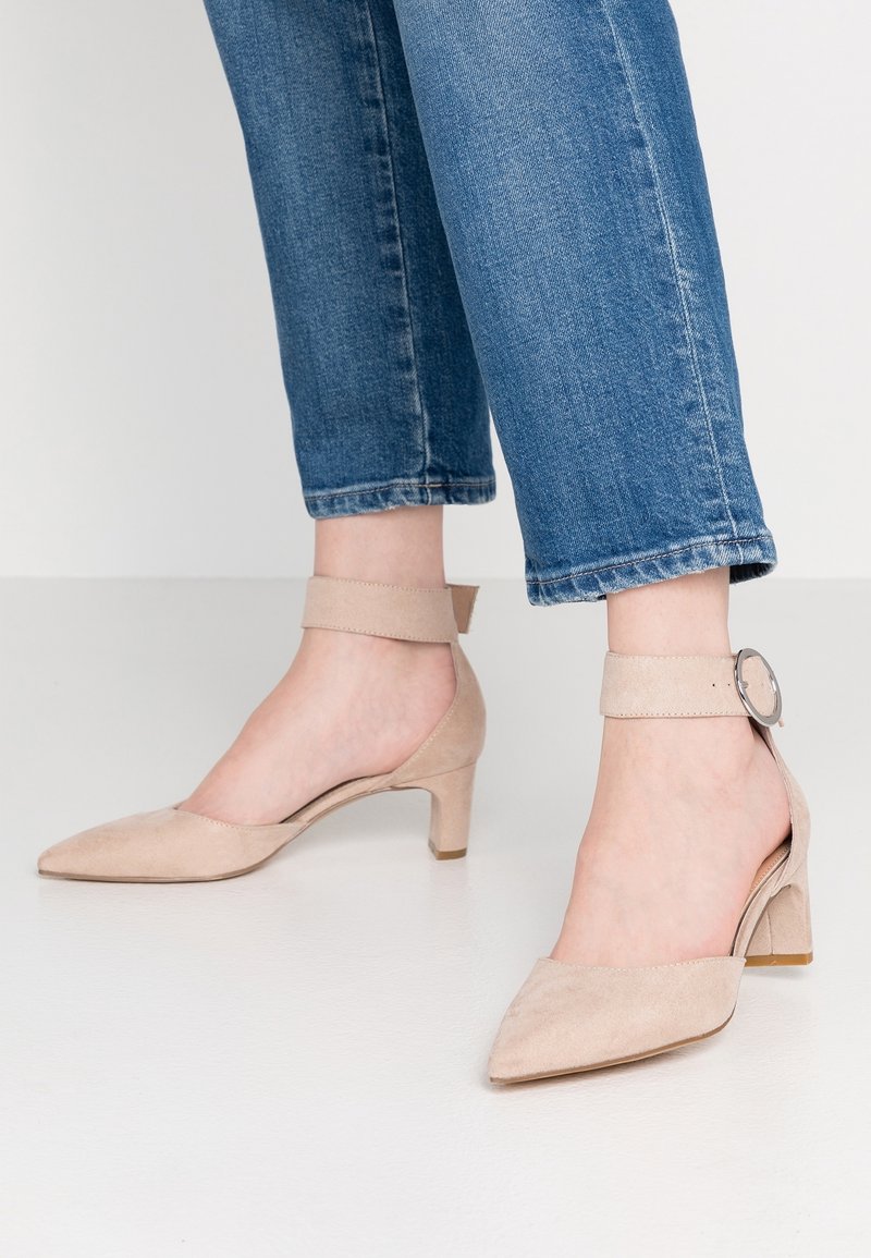 Talons à bride de cheville en daim beige avec un bout pointu et un talon bloc modeste. La bride présente un détail de boucle en métal rond.