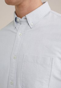 Chemise bleu clair à manches longues avec col étalé, poche avant et boutons blancs. Le tissu a une texture lisse avec un léger motif tissé.
