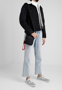 Giacca di jeans nera con colletto in shearling bianco, maglione dolcevita bianco, jeans dritti azzurri e sneakers grigie, mentre tiene una piccola borsa nera.