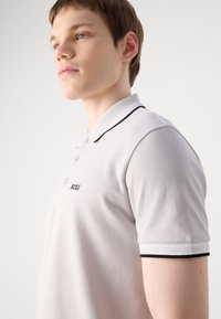 Polo cinza claro com detalhes em preto, apresentando uma pala de três botões e o logótipo "BOSS" bordado no peito. Textura suave.