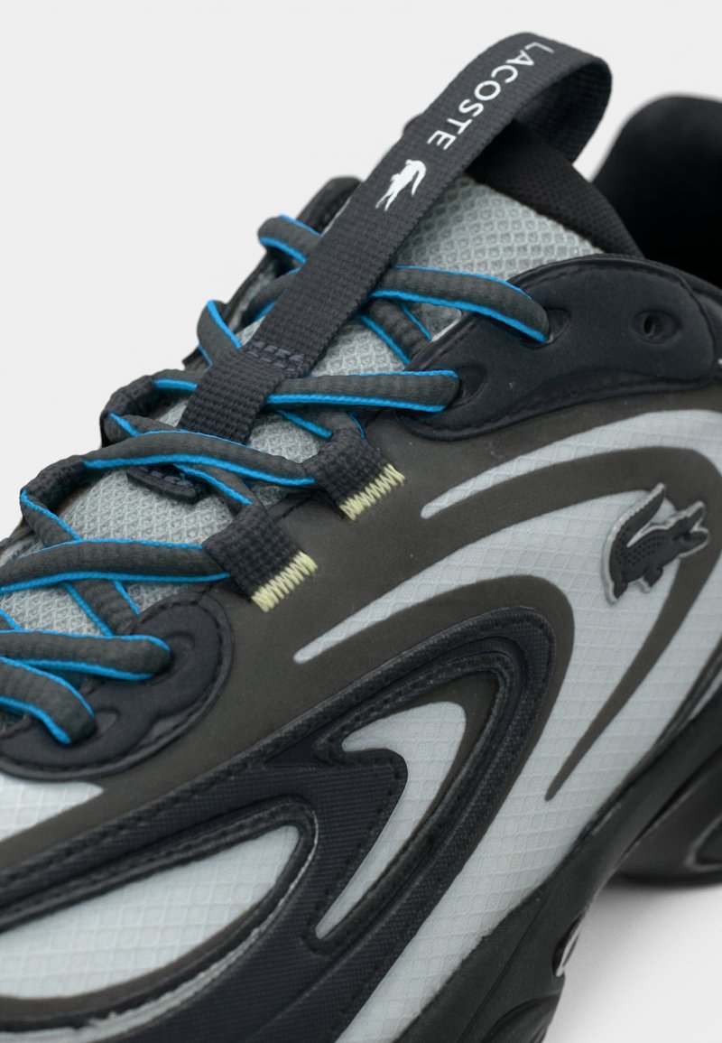 Chaussure de sport grise et noire avec une tige en mesh texturé, des accents bleus et un design incurvé distinctif avec un logo bien visible.