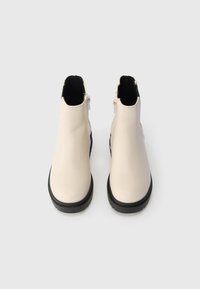 Beiget nilkkurit, joissa on sileä pinta, musta pohja ja sivussa vetoketju helppoa pukeutumista varten. Kaksivärinen muotoilu, jossa laivastonsinisiä yksityiskohtia.