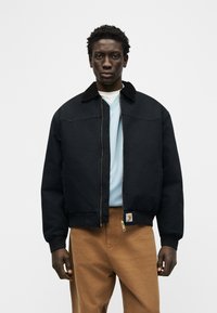 Άνδρας μοντέλο φορούντας μαύρο τζάκετ με φερμουάρ πάνω από γαλάζιο πουκάμισο και καφέ παντελόνι, στέκεται μπροστά σε απλό λευκό φόντο.