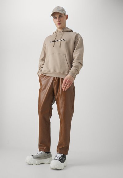 Sudadera beige con un bolsillo frontal y texto gráfico, combinada con pantalones marrón de aspecto cuero y zapatillas gruesas blancas y grises.