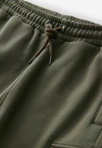 Olivgrüne Sweatpants verfügen über einen elastischen Bund mit einem Kordelzug, Metall-Ösen und einem strukturierten Stoff. Praktische Seitentaschen sind ebenfalls vorhanden.