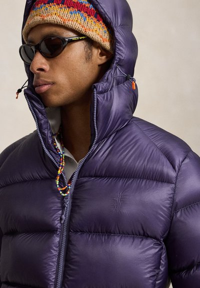 Man wearing a colorful knit beanie, black sunglasses with Polo Sport logo, and a zipped purple puffer jacket with hood and beaded necklace.
