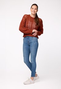 Roestkleurige gebreide trui met een gestructureerd, open steekpatroon, gecombineerd met lichtblauwe skinny jeans met versleten details en witte sneakers.