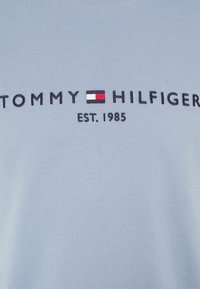 Camiseta de cuello redondo en azul claro, hecha de un tejido suave, que presenta el logo bordado "TOMMY HILFIGER" y "EST. 1985" en azul marino.