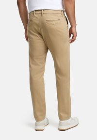 Pantalon chino en coton beige avec une coupe ajustée, poches arrière boutonnées et une texture lisse. Associé à des baskets blanches pour un contraste.