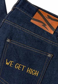Pantaloni in denim blu scuro con ricami a contrasto in oro che recitano "WE GET HIGH" sulla tasca posteriore, con un'etichetta in pelle e dettagli in rivetti metallici.