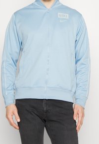 Giacca Nike azzurro chiaro con zip, colletto alto, tasche laterali, strisce bianche decorative e logo sul lato superiore sinistro. Tessuto dalla texture liscia.