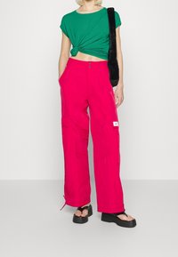 Růžové oversized kalhoty s volným střihem, se strukturovaným povrchem a cargo kapsami, doplněné zeleným crop topem s uzlem. Černé platformové sandály.