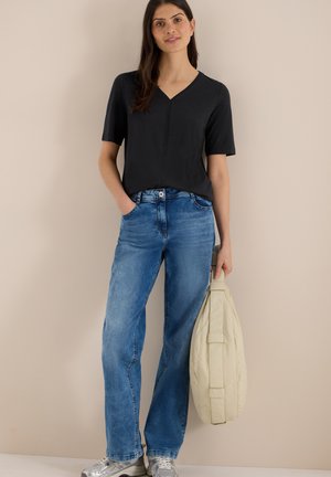 Frau trägt schwarzes V-Ausschnitt-T-Shirt mit kurzen Ärmeln, blaue Jeans, silberne Turnschuhe, hält eine beige gepolsterte Tasche vor einem einfarbigen beigen Hintergrund.
