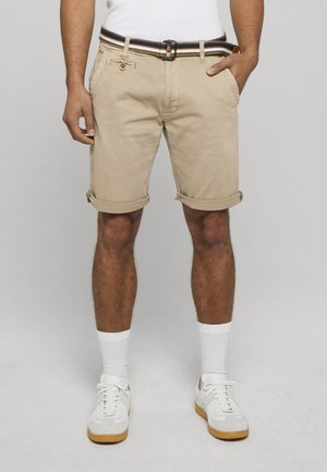Mann iført beige opprullede shorts med et stripete belte, hvite sokker og hvite joggesko, står mot en grå bakgrunn.