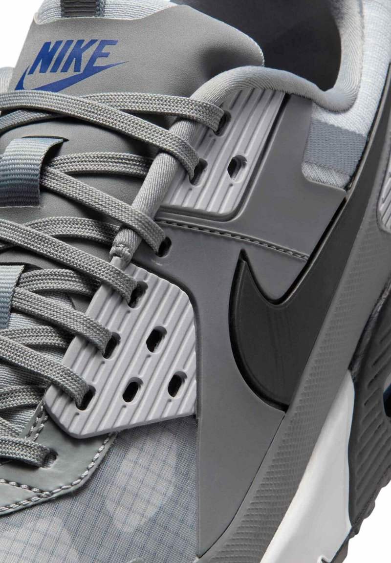 Gros plan sur une basket Nike grise avec logo bleu, swoosh noir, lacets texturés et panneau d'œillets en plastique côtelé.