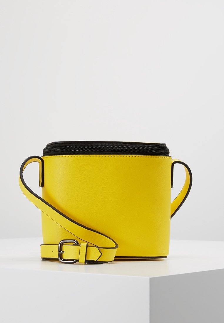 Sac bandoulière en cuir jaune avec fermeture éclair noire et sangle ajustable, présenté sur une surface blanche contre un fond clair uni.