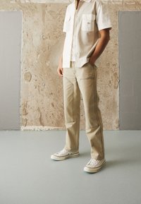 Camicia beige di cotone con bottoni e due tasche sul petto indossata sopra una t-shirt bianca; abbinata a pantaloni beige con doppie cuciture e sneakers bianche.