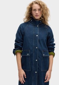 Niebieska kurtka dżinsowa z wysokim kołnierzem, zatrzaskami, dwoma kieszeniami z przodu i podwiniętymi rękawami, odsłaniającymi kontrastujące zielone wnętrze.