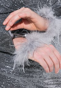 Stříbrné flitrové šaty s průhlednými šedými peřím na manžetách; viditelný detail zapínání na zápěstí ruky, která drží manžetu.