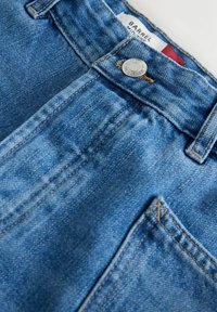 Jasnoniebieskie dżinsy z widocznym przeszyciem, metalicznym zapięciem na guzik oraz metką z napisem „BARREL” w pobliżu talii.