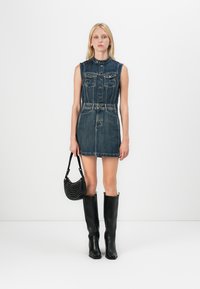 Denim sleeveless dress with front button closure, two chest pockets, belted waist, and a flared hem. Paired with knee-high black boots and a black bag.